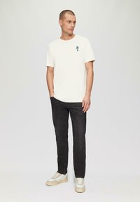 Korte mouw crèmekleurig t-shirt met een kleine blauwe graphic, gecombineerd met donkere jeans en witte sneakers met beige zolen.