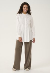 Camicia bianca con bottoni e maniche lunghe, dotata di un colletto arricciato, abbinata a pantaloni ampi a righe marroni e scarpe di colore chiaro.