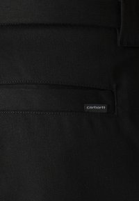 Crni materijal s dijagonalnom teksturom; sadrži zakrpatnu džep i vidljivu tagu Carhartt u sivoj boji s desne strane.
