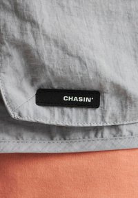 Tessuto grigio con texture stropicciata presenta una etichetta nera CHASIN'. L'indumento è parzialmente mostrato con un tessuto di colore corallo sotto.