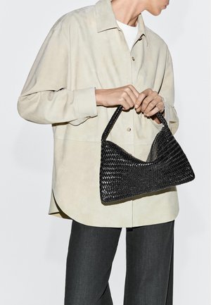 Person trägt beige Hemd und schwarze Hose und hält eine schwarze, gewebte Handtasche mit kurzem Henkel auf Taillenhöhe.