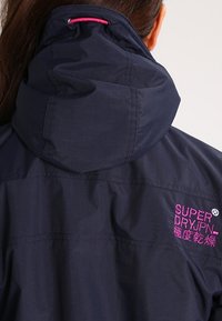 Mørkeblå hættetrøje med tekstureret stof. Har lyserødt broderet logo på ryggen og snøre til justerbar pasform.