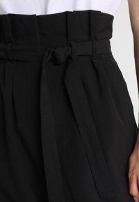 Pantalons noirs taille haute avec une large ceinture, une ceinture en tissu avec un nœud, et un design plissé. Tissu lisse et texturé.