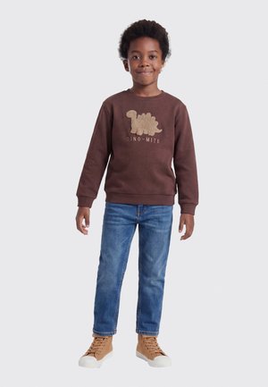 Dreng stående iført brun dinosaur-sweatshirt, blå jeans og beige høje sneakers på ensartet lyst baggrund.