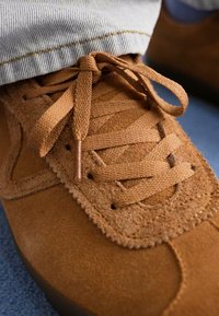Sapatilhas de camurça castanha com atacadores combinando atados em laço, usadas com jeans de tom claro contra um fundo desfocado.