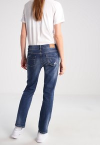 Jeans de mezclilla azul con corte Slim, cinco bolsillos y un ligero desgaste en los bolsillos traseros, combinados con una camiseta blanca de cuello redondo y zapatillas blancas.