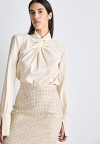 Cremefarvet bluse med snoet halsudskæring, lange ærmer, og en lys beige, struktureret blyantskirt med et vævet mønster og subtile metalliske detaljer.