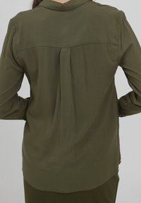 Olivengrön långärmad skjorta, lättviktsmaterial, spetsig krage, detalj med veck bak, slät textur, rundad fåll.