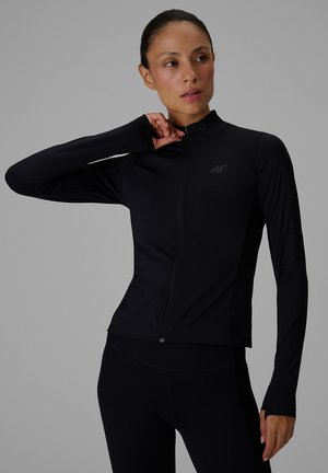 Femme en veste de sport noire à manches longues zippée et leggings, ajustant le col d'une main, regardant légèrement sur le côté sur un fond uni.