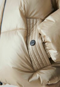 Poche de veste matelassée beige avec des détails cousus et un bouton rond noir étiqueté "Clarks."