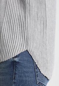 Hellgrauer gerippter Pullover mit V-förmigem Saum, strukturiertem Stoff, geschichtetem Design, kombiniert mit blauen Jeans.