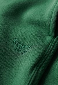 Zielona dzianina z haftowanym logo "Superdry" w nieco ciemniejszym odcieniu zieleni. Miękka tekstura z zbliżeniem na szwy.
