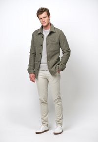 Olivgrön ullblandning jacka med svarta knappar, framficka; grå stickad tröja under, ljusbeige byxor och vita sneakers.