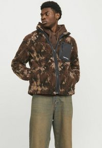 Brown fuzzy jacket with a black zippered pocket, featuring a camouflage pattern, high collar, and a soft texture. Worn over a brown hoodie.