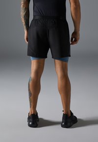 Svart idrettsshorts med elastisk midje, kombinert med lyseblå kompresjonsshorts underneath. Svart idrettssko og en tatovering synlig på beinet.