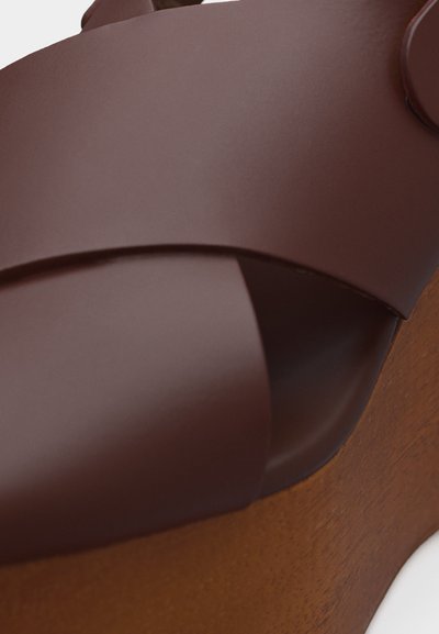 Gros plan sur une sandale en cuir marron avec des larges brides qui se chevauchent et un talon compensé en bois, posée sur une surface blanche.