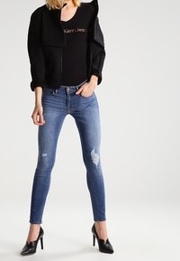 Chaqueta negra corta, camiseta negra ajustada, jeans skinny azules desgastados y tacones altos negros. Los vaqueros presentan desgarros y un lavado medio.