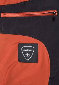 Pomarańczowo-czarna część kurtki z zamykaną na zamek kieszenią oraz naszywką z logo Strellson w kształcie trójkąta z białymi detalami.