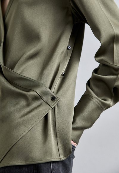 Olivgrüne Bluse mit seidiger Textur, die über einen Knopfverschluss vorne, einen schrägen Saum und seitliche Klappendetails verfügt.