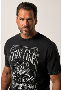 Camiseta de algodón negra con una impresión gráfica que incluye las palabras "Alimenta el fuego, alimenta el alma" y dibujos de llamas y utensilios de cocina.