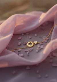 Bracciale d'oro con chiusura a forma di cuore e un accento decorativo scintillante a forma di cuore, adagiato su un tessuto rosa e piccole pietre viola.
