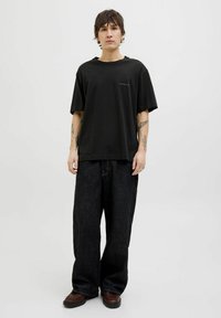 T-shirt noir oversize avec un col rond et un petit détail en texte bleu. Associé à un jean large foncé et des chaussures marron. Style simple et décontracté.
