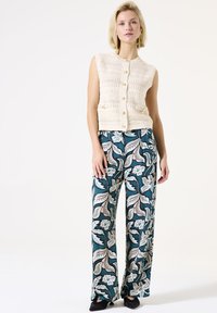 Gilet en crochet crème avec des boutons sur le devant, associé à un pantalon large teal présentant un motif floral en blanc et rouge. Chaussures noires visibles.