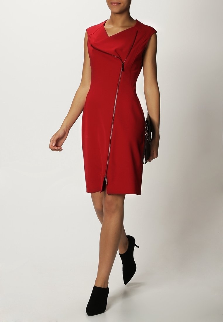 Femme marchant portant une robe rouge sans manches avec une fermeture éclair diagonale et des bottines noires à talons, tenant une pochette noire.