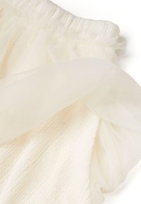 Tejido blanco texturizado con patrones de punto y un suave tul transparente arrugado en la cintura. Apariencia ligera y etérea.