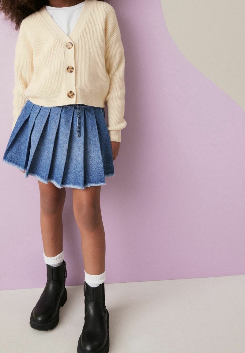 Enfant portant un cardigan crème boutonné, une chemise blanche, une jupe en jean plissée, des chaussettes blanches et des bottines noires, debout devant un mur rose et beige.