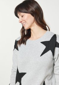 Pulover gri tricotat cu modele mari de stele negre, guler rotund și mansete cu nervuri; textura moale adaugă confort.