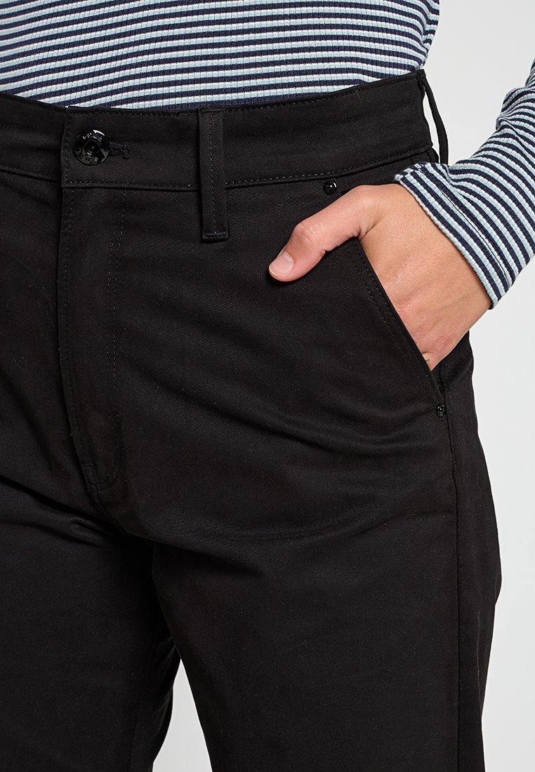 Un pantalon en denim noir présente une poche avant, une fermeture à bouton et des passants de ceinture. Une chemise à rayures à manches longues est partiellement visible au-dessus.