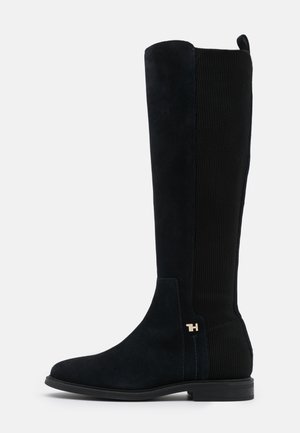 Botte noire montante jusqu’au genou avec devant en daim, dos en tissu côtelé, talon bas et petit logo doré "TH" près de la cheville sur fond blanc.