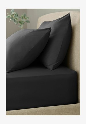Zwei schwarze Kissen, die auf einem schwarzen Spannbettlaken auf einem beigen gepolsterten Bettrahmen mit einer grünen Pflanze im Hintergrund liegen.