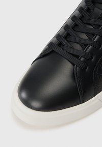 Sneaker din piele neagră cu un finisaj neted, vârf rotunjit și talpă din cauciuc albă. Are șireturi negre și un design subtil embosat.