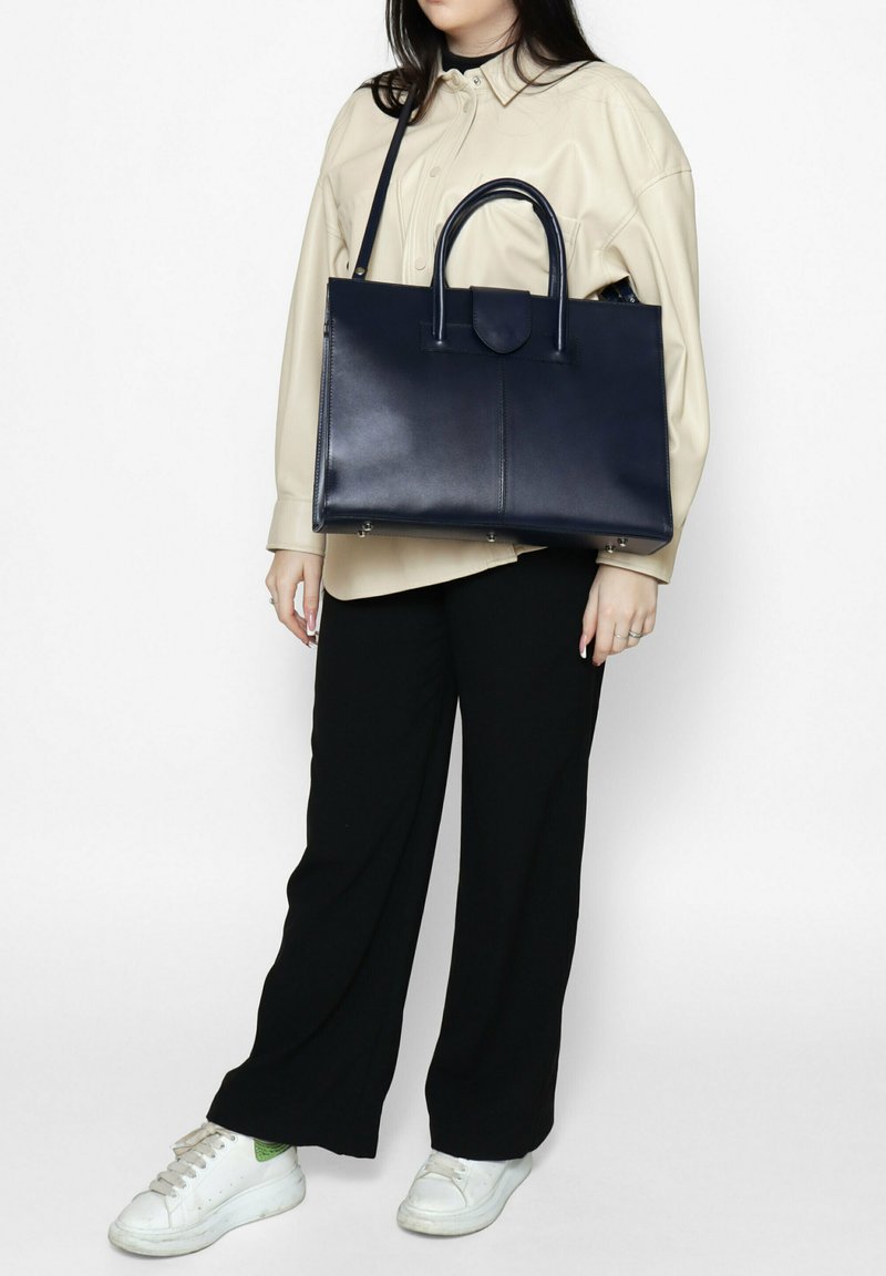 Sac fourre-tout en cuir marine avec deux poignées supérieures, une fermeture magnétique et une finition lisse. Le sac présente une forme rectangulaire et des pieds en métal.