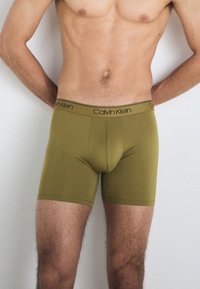 Alyvuogių spalvos Calvin Klein bokseriai su juodu logotipu, pasižymintys lygiu paviršiumi ir prigludusia forma, pasiekiančia iki šlaunies vidurio.