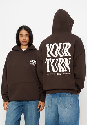 Deux personnes portant des sweat-shirts à capuche marron foncé et des jeans bleus, l’une face à l’avant et l’autre de dos, montrant le texte "YOUR TURN" sur le sweat.