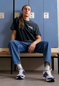 Schwarzes T-Shirt mit dem Text "INTER", hellblaue Jeans und weiße Sneakers mit lila Akzenten; sitzt auf einer Holzbank in einem Umkleidebereich.