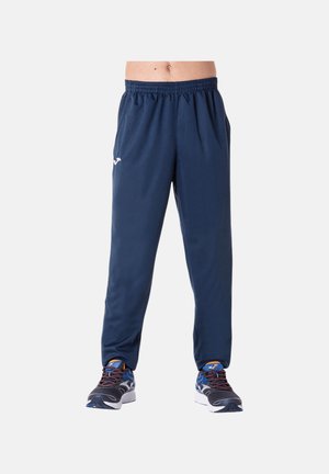 La persona lleva pantalones de chándal azul marino y zapatillas negras y azules, de pie contra un fondo blanco liso.