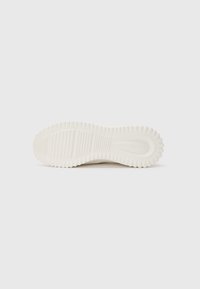 Suela de zapatilla de goma blanca con textura estriada, que presenta una forma redondeada y un sutil estampado de logo. Elementos de diseño limpios y mínimos.