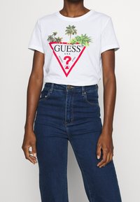 Bílý bavlněný tričko s barevným grafickým motivem palmy a červeným trojúhelníkovým logem, v kombinaci s tmavě modrými džíny s vysokým pasem.