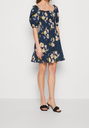 Femme portant une robe courte bleu marine à motifs floraux avec des manches bouffantes et des sandales à talons noires, debout devant un fond clair uni.