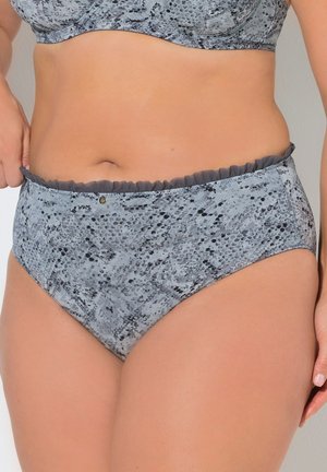 Mujer ajustándose ropa interior gris con estampado de leopardo y cintura alta con volantes, lleva un sujetador a juego, torso visible sobre fondo liso.