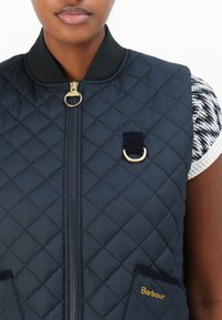 Colete acolchoado azul-marinho com gola redonda, fecho dourado e bolsos frontais. Apresenta um detalhe do logo dourado e um acento em laço próximo ao zíper.