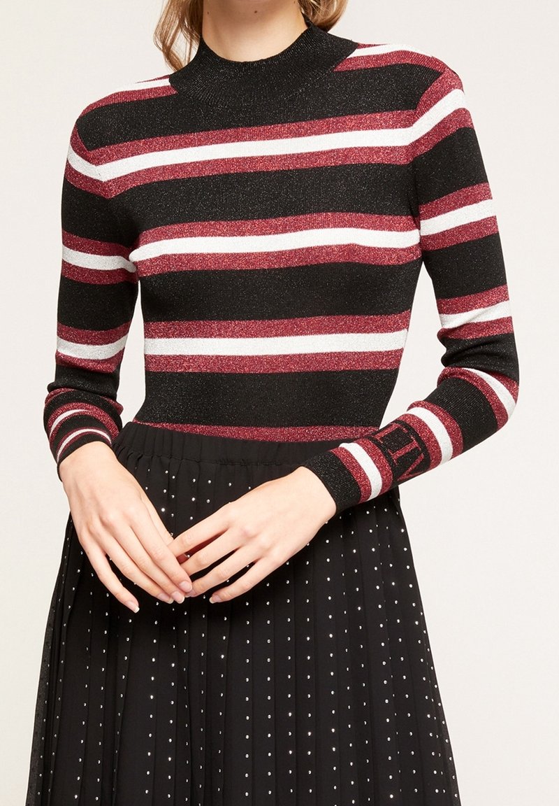 Femme portant un haut à manches longues rayé noir, rouge et blanc, associé à une jupe plissée noire ornée de petits pois blancs.