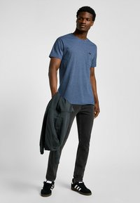 Blauwe heather t-shirt met korte mouwen en een borstzak, gecombineerd met grijze slim-fit jeans en zwarte sneakers met witte en grijze accenten.