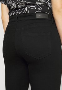 Jeans negros con un diseño ajustado, textura suave y dos bolsillos traseros con detalles de costura sutil. Se puede ver una etiqueta de la marca en la cintura.