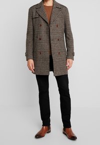 Abrigo de cuadros marrones de doble pecho con grandes botones, llevado sobre un suéter marrón y combinado con pantalones negros y botas de tobillo marrones.