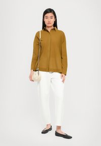 Camicia a maniche lunghe color senape, pantaloni bianchi a sigaretta, ballerine nere e una piccola borsa beige con tracolla a catena. Tessuto liscio, design aderente.